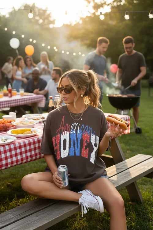 Southern Charm Design Works American Honey graphic t-shirt in brown with distressed red, white, and blue lettering. Model wears it at a picnic.